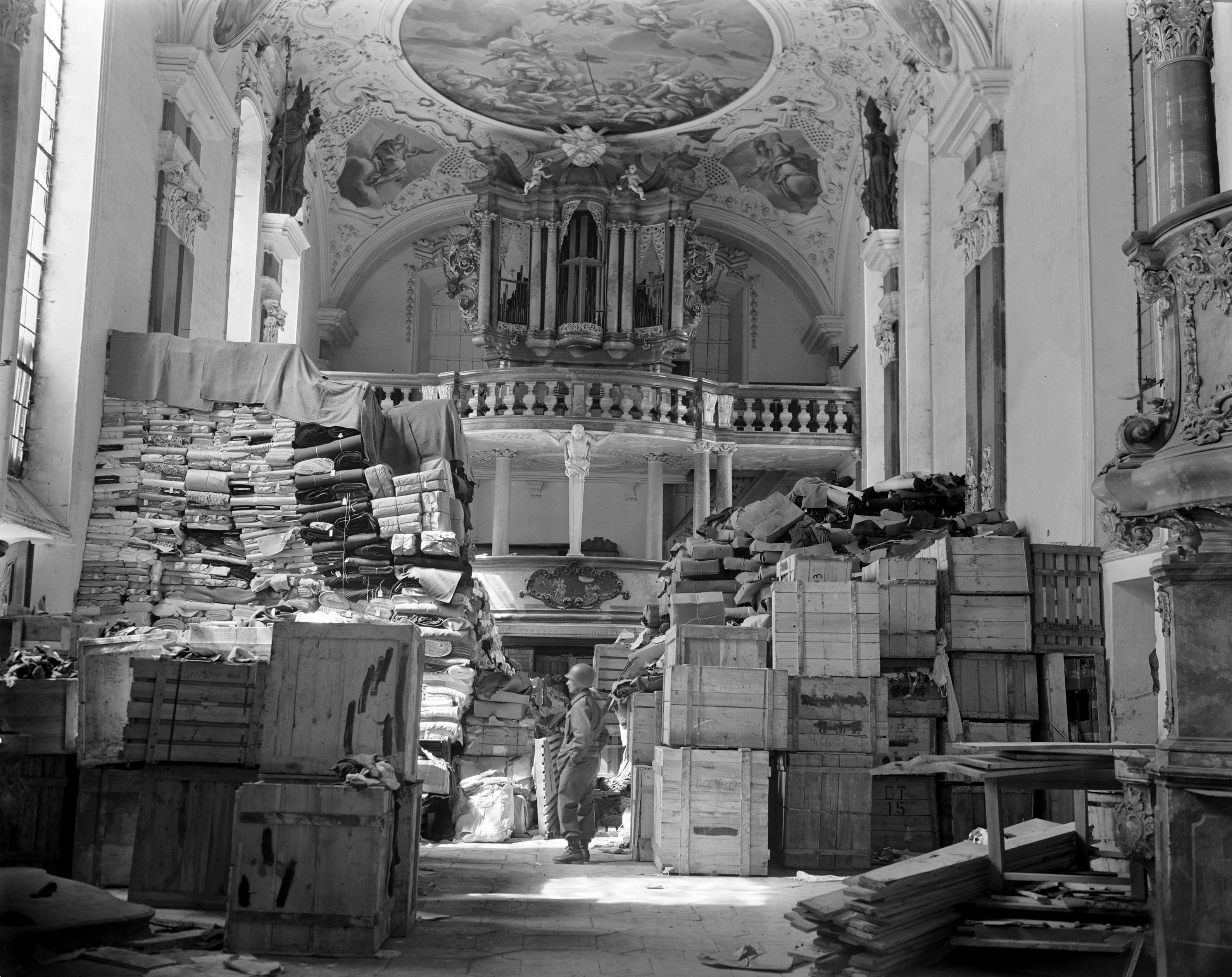 Veduta interna in bianco e nero di una chiesa barocca a Ellingen, in Germania, colma di oggetti saccheggiati appartenuti a vittime ebree dell’Olocausto. Tra balle di tessuti, casse e pacchi impilati si trova un soldato statunitense. Fotografia del 24 aprile 1945.