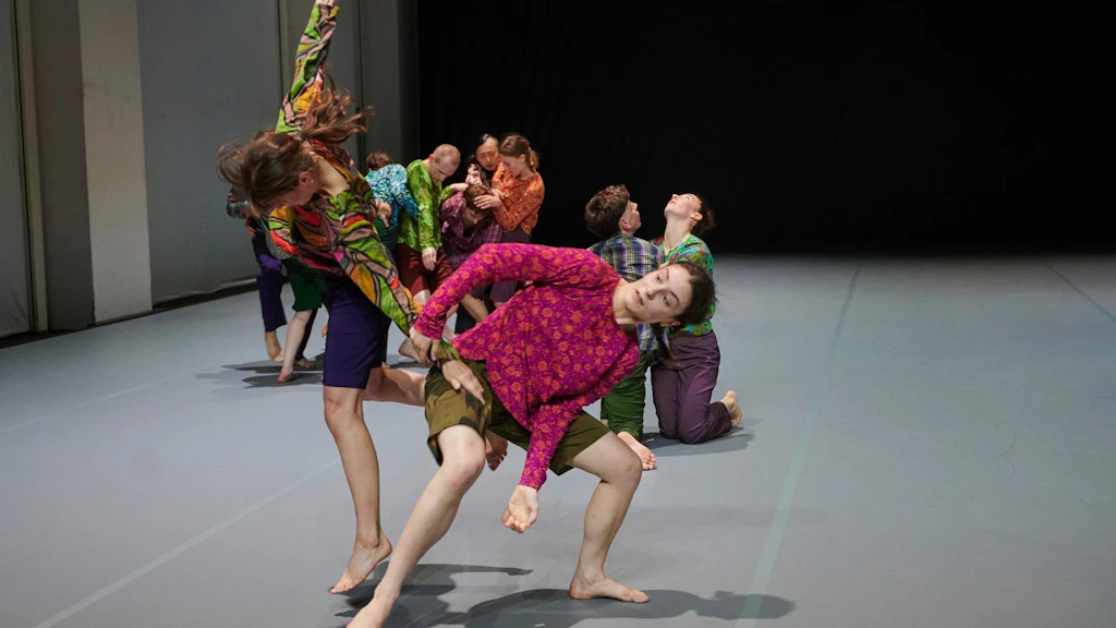 Scena di danza contemporanea con più danzatrici e danzatori in costumi colorati che eseguono movimenti dinamici e intrecciati sul palcoscenico.