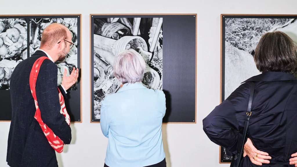 Tre persone osservano opere in bianco e nero in una mostra. Le immagini raffigurano strutture e oggetti dettagliati, incorniciati su una parete bianca.