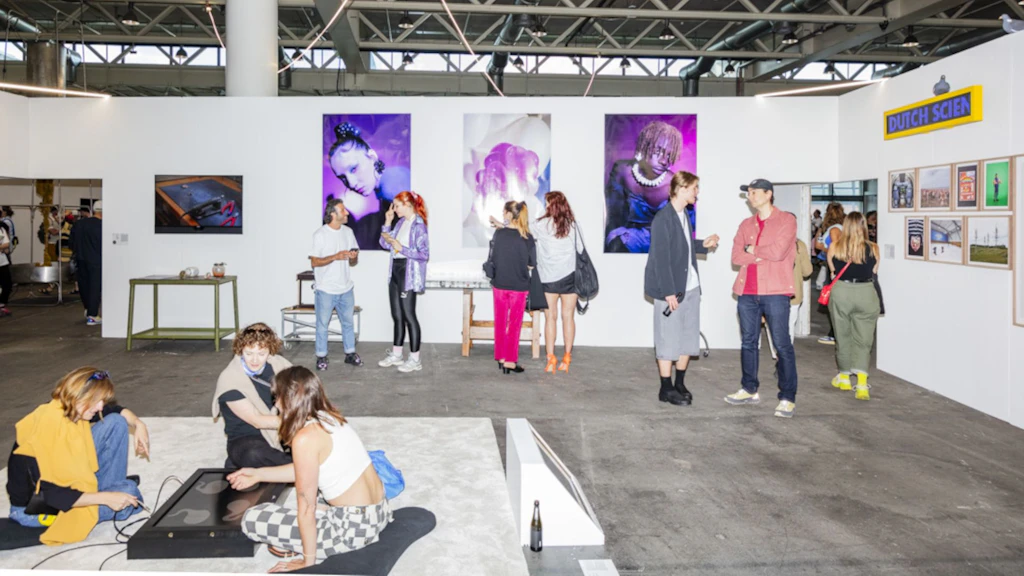 Mostra di design con più visitatori che osservano opere d’arte. Alle pareti sono appesi ritratti e manifesti dai colori vivaci, mentre in primo piano alcune persone sedute su una piattaforma interagiscono con un’installazione.