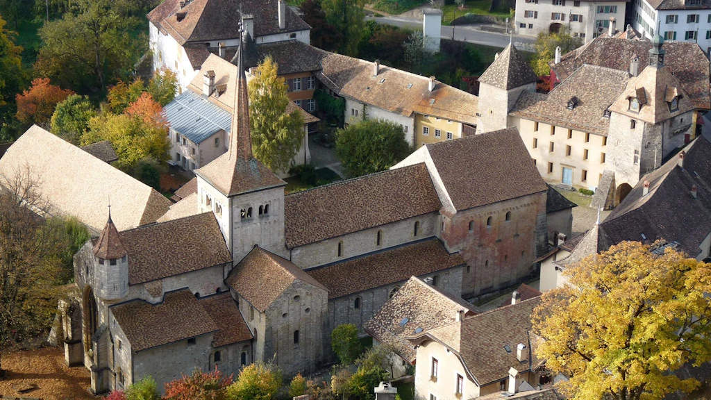 Veduta aerea del villaggio di Romainmôtier e della sua chiesa abbaziale cluniacense, circondati da edifici storici, alberi autunnali e tetti in tegole rosse.