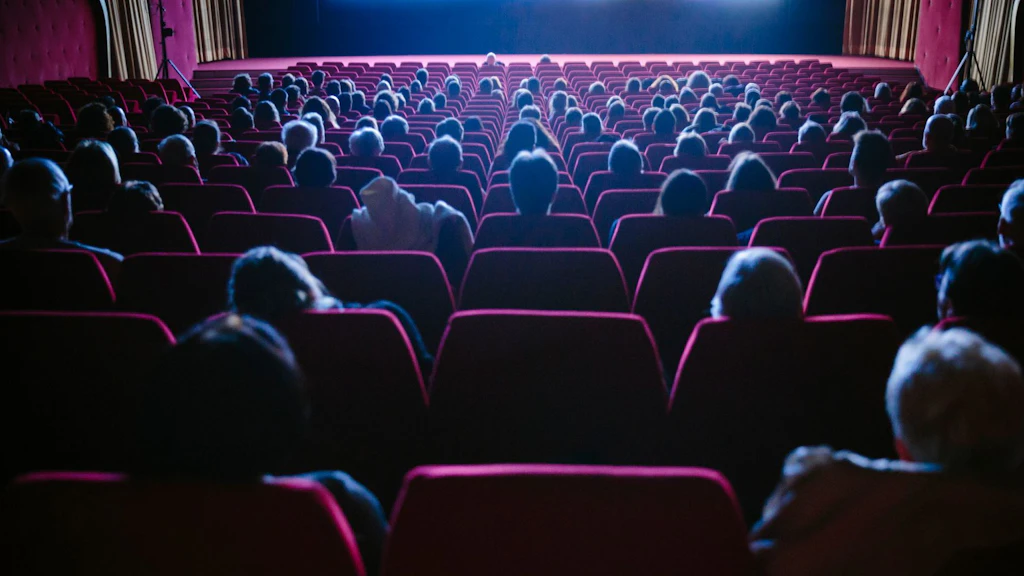 Salle de cinéma avec fauteuils bordeaux et rideaux jaune foncé vue depuis le public. Les spectateurs regardent vers l’avant en direction de l’écran lumineux.