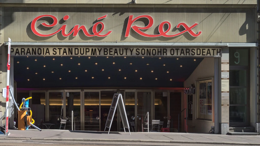 Façade du cinéma Rex à Berne avec l’inscription rouge « Ciné Rex », une entrée éclairée et une bande annonçant des titres de films au-dessus des portes vitrées.