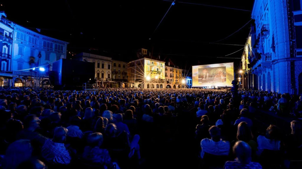 Un large public réuni sur la Piazza Grande lors du Festival du film de Locarno assiste le soir à une projection en plein air sur un grand écran, entouré de bâtiments historiques et éclairé par des projecteurs.
