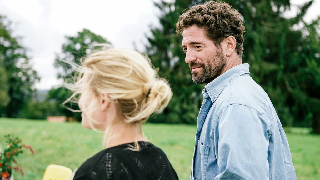 Scène du film « Le vent tourne » de Bettina Oberli : une femme et un homme se tiennent dans une prairie devant un paysage rural ; l’homme regarde la femme tandis qu’elle contemple le paysage.