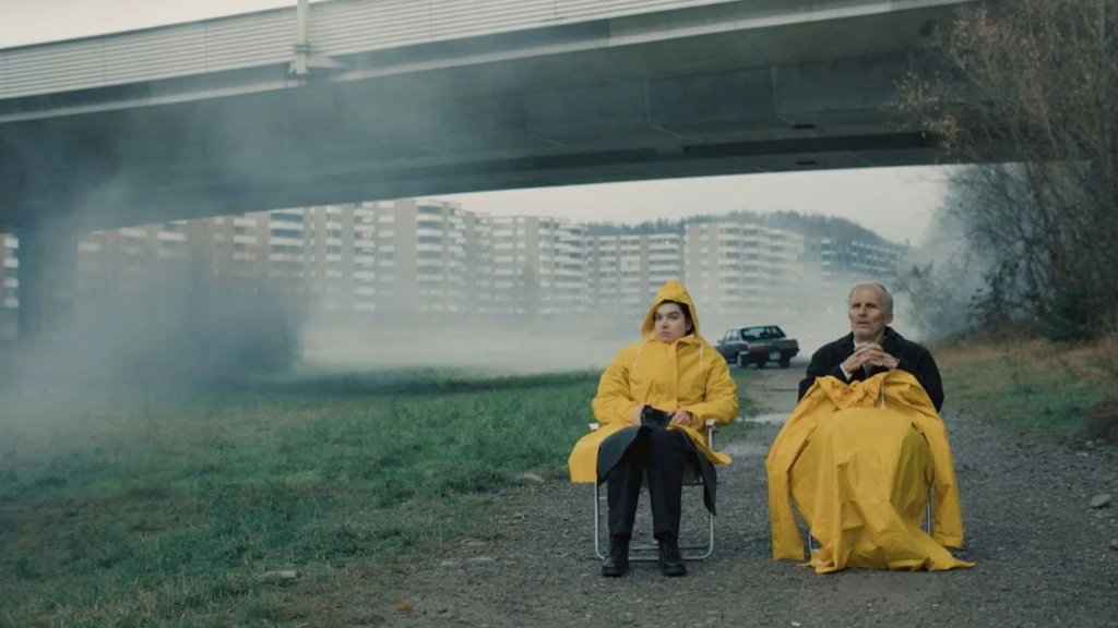 Deux personnes en imperméables jaunes sont assises sur des chaises pliantes sous un pont autoroutier. L’environnement brumeux et gris ainsi que le paysage désert créent une atmosphère sombre.