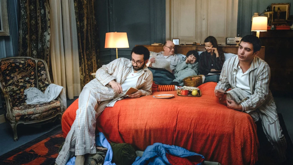 Plusieurs personnes en tenue de nuit sont assises et allongées dans une chambre sur un grand lit couvert d’une couverture rouge. La scène familiale paraît calme, tandis que certaines personnes lisent, discutent ou regardent attentivement dans une même direction.
