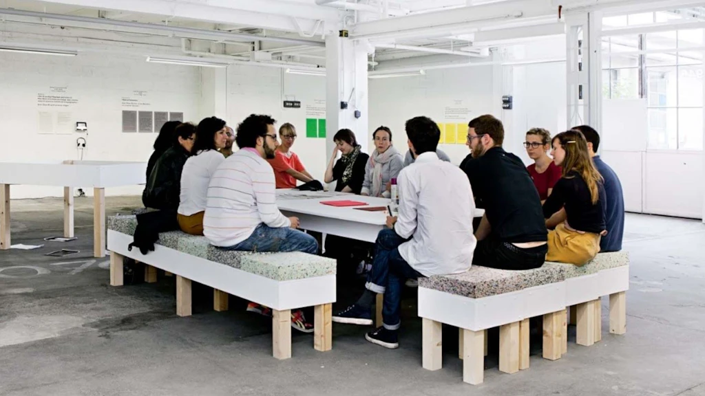 Plusieurs personnes sont assises autour d’une grande table dans un espace d’exposition lumineux et discutent. L’environnement ouvert, avec des panneaux de texte et des installations, montre un échange commun.