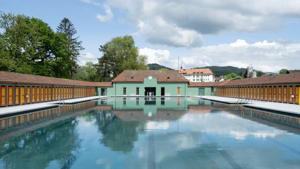 Ein grosses, rechteckiges Freibad mit klarem Wasser, umgeben von langen Reihen gelb-orangefarbiger Umkleidekabinen und einem zentralen, mintgrünen Gebäude; im Hintergrund Bäume, Hügel und einige Stadtgebäude unter leicht bewölktem Himmel.