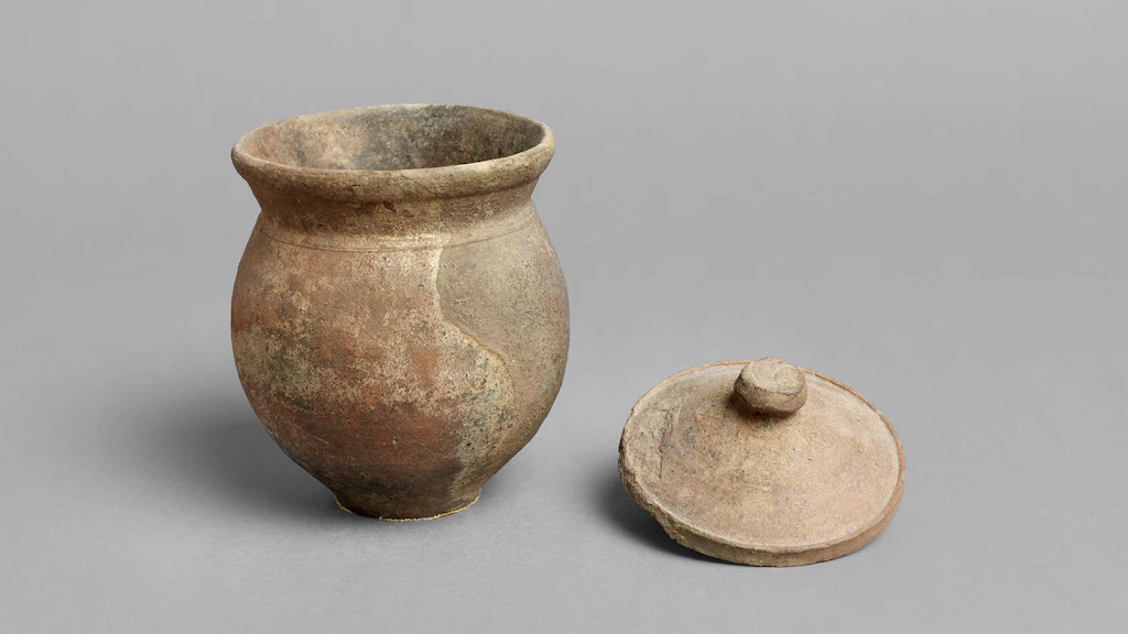 Kleine Vase aus Ton («Olletta») mit abnehmbarem Deckel vor grauem Hintergrund. Das Gefäss aus dem 3.–1. Jahrhundert v. Chr. weist Gebrauchsspuren und unregelmässige Verfärbungen auf.