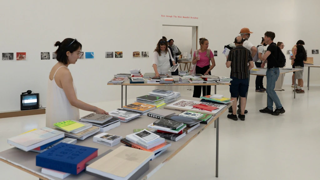 Heller Ausstellungsraum mit mehreren Besucherinnen und Besuchern, die Bücher der Ausstellung «Die schönsten Schweizer Bücher» betrachten. Auf Tischen sind zahlreiche Publikationen ausgelegt, während sich die Gäste durch den Raum bewegen und austauschen.