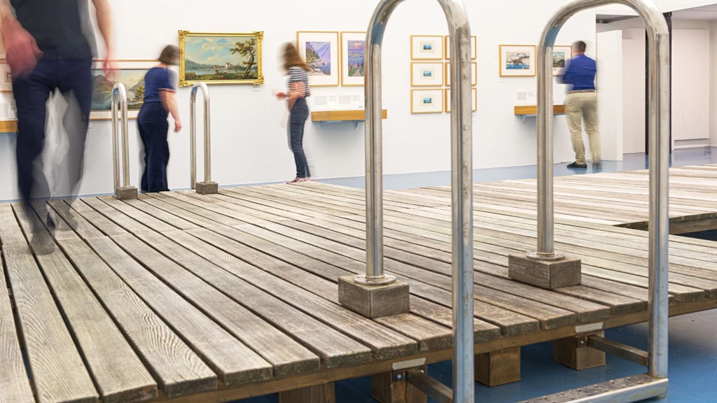 Besucherinnen und Besucher bewegen sich in der Ausstellung «Seen, Spiegel der Schweiz» in der Schweizerischen Nationalbibliothek, mit begehbarer Steg-Installation und gerahmten Bildern an den Wänden.
