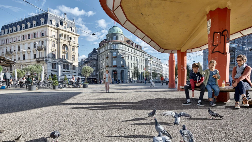 Zentralplatz in Biel: Im Hintergrund fassen Gebäudefassaden den Platz ein und es gibt ein Restaurant mit Aussenbestuhlung. Im Vordergrund sitzen eine Frau und zwei Kinder in einer Tramhaltestelle mit Bank, roten Stützen und weitläufiger Überdachung. Sie füttern Tauben. 