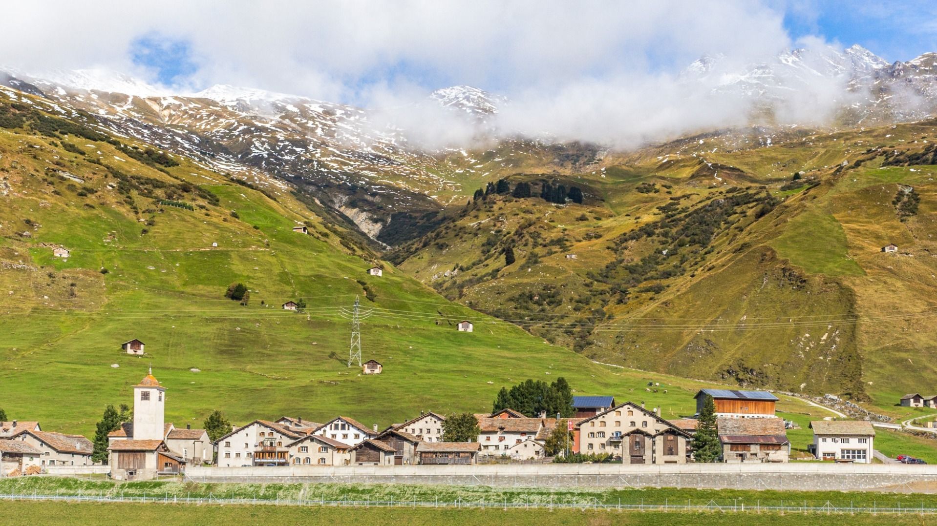 Ein kleines Dorf liegt am Fuss grüner Berghänge mit verstreuten Hütten und schneebedeckten Gipfeln im Hintergrund. Die Wolken hängen tief über den Bergen.
