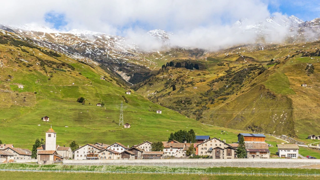 Ein kleines Dorf liegt am Fuss grüner Berghänge mit verstreuten Hütten und schneebedeckten Gipfeln im Hintergrund. Die Wolken hängen tief über den Bergen.