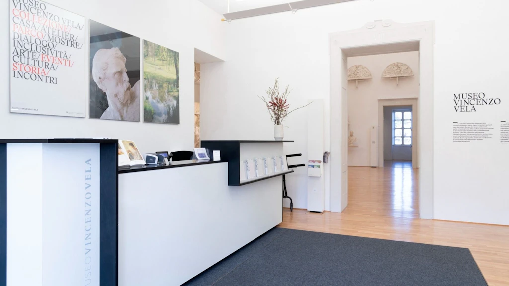 Eingangsbereich des Museo Vincenzo Vela mit Empfangstheke, Informationsmaterial und Wandbildern; Blick in die dahinterliegenden Ausstellungsräume des historischen Künstlerhauses.