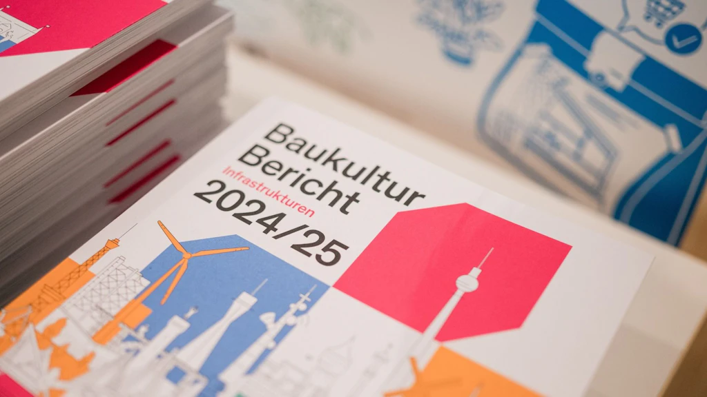 A stack of the Baukultur Report 2024/25 with graphic illustrations of infrastructures on the cover. The book is on a table, with more brochures visible in the background.