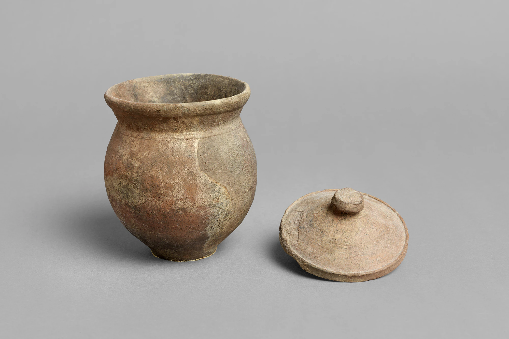 Petite jarre en terre cuite (« olletta ») avec couvercle amovible sur fond gris. Le récipient, datant du IIIe au Ier siècle av. J.-C., présente des traces d’usage et des décolorations irrégulières.