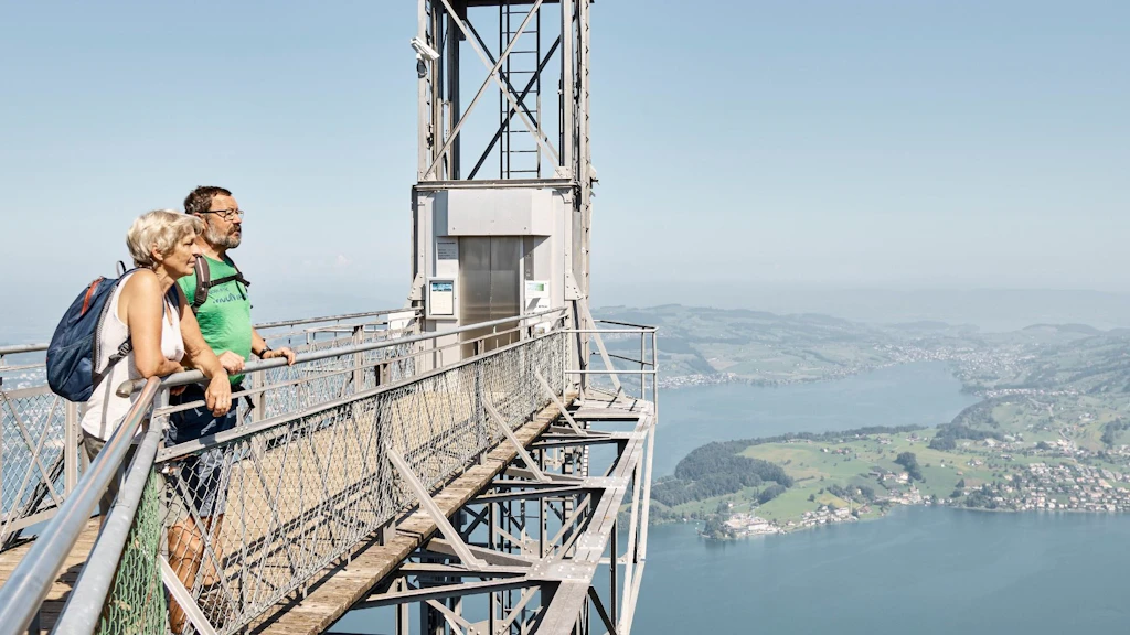 Un homme et une femme en vêtements de randonnée admirent le paysage offert par le lac des Quatre-Cantons, perchés en toute sécurité au-dessus du vide sur la plateforme d’accès de la station supérieure de l’ascenseur du Hammestschwand.