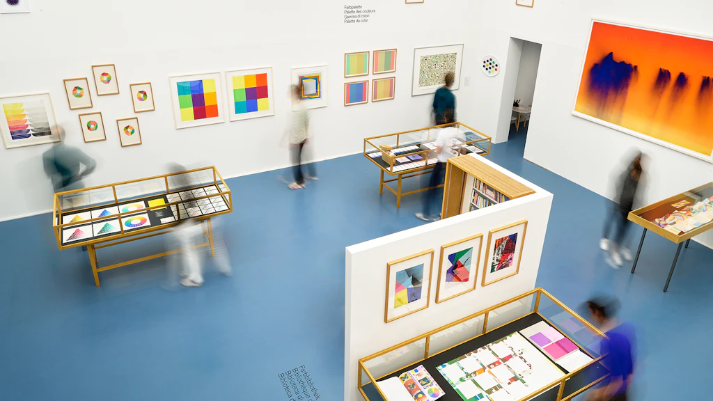Vue d’une salle d’exposition lumineuse avec des vitrines, des œuvres imprimées et plusieurs visiteurs en mouvement. L’exposition « Espace de couleur » présente des compositions chromatiques et des œuvres graphiques sur des murs blancs et un sol bleu.