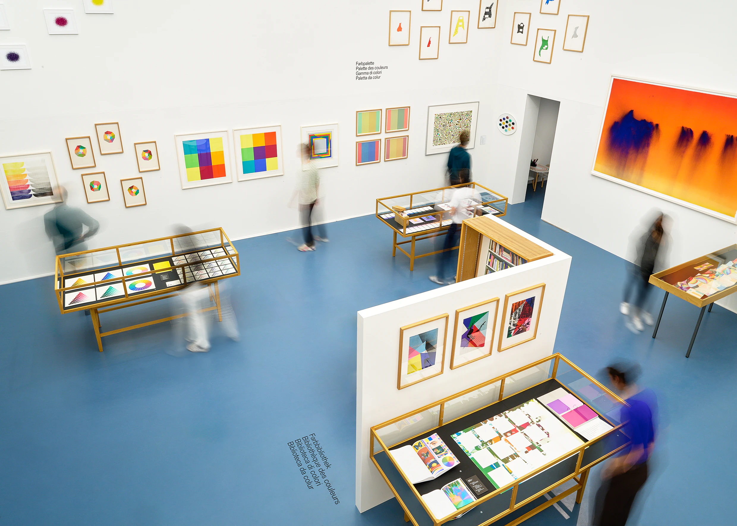 Vue d’une salle d’exposition lumineuse avec des vitrines, des œuvres imprimées et plusieurs visiteurs en mouvement. L’exposition « Espace de couleur » présente des compositions chromatiques et des œuvres graphiques sur des murs blancs et un sol bleu.