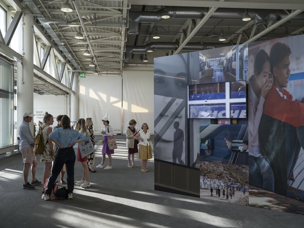 Des visiteurs se tiennent dans une halle d’exposition à côté d’une installation grand format composée de photographies et d’écrans.
