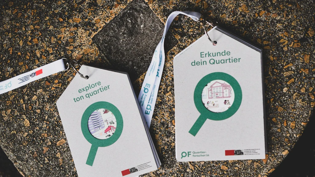 Deux étiquettes en carton en forme de maison avec des rubans blancs sont posées sur une pierre. La carte de gauche porte le texte « explore ton quartier », celle de droite « Erkunde dein Quartier ». Toutes deux affichent un symbole de loupe verte avec de petites illustrations de bâtiments au centre.