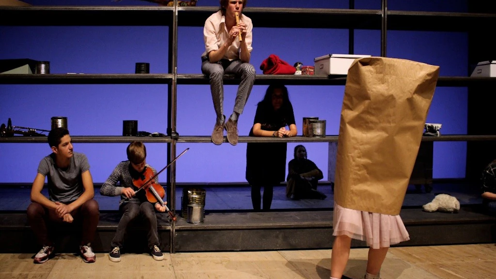 Des jeunes se tiennent sur une scène de théâtre ; une personne joue de la flûte à bec, d’autres sont assises sur une structure du décor.
