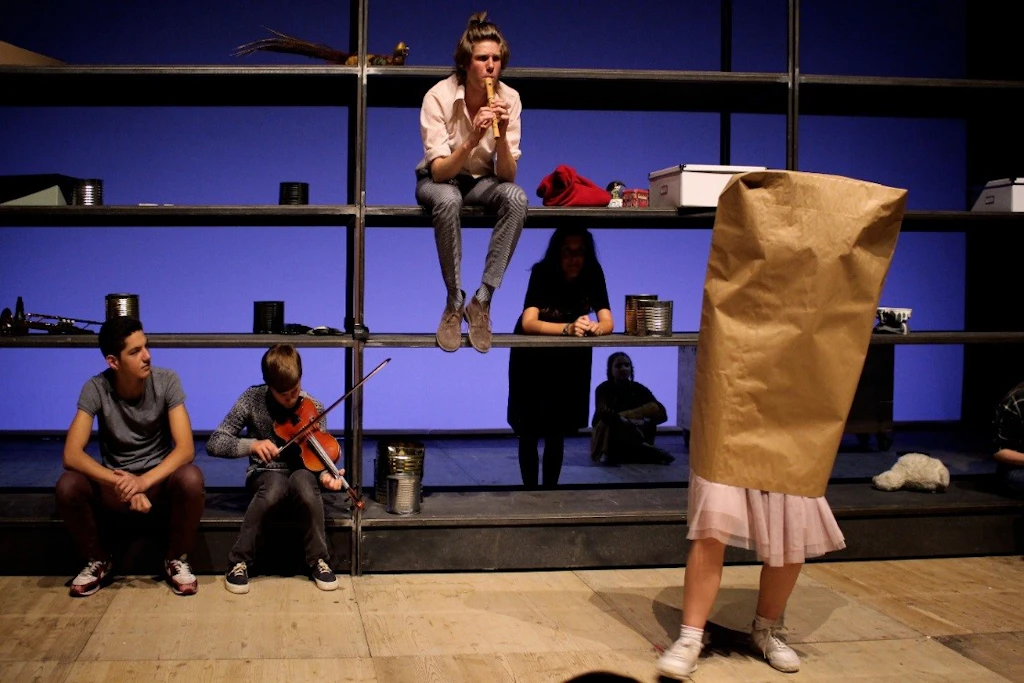 Des jeunes se tiennent sur une scène de théâtre ; une personne joue de la flûte à bec, d’autres sont assises sur une structure du décor.