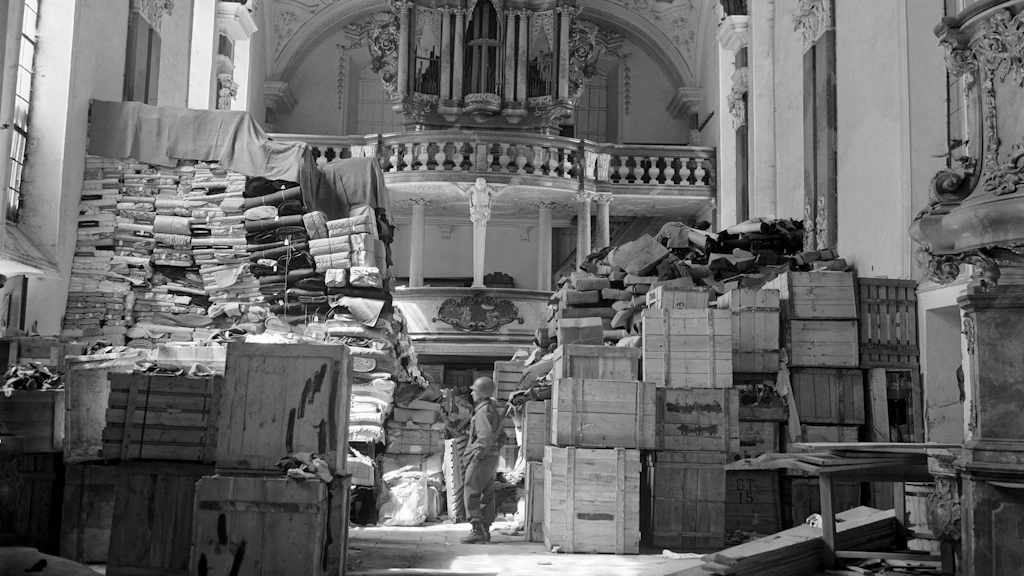 Vue intérieure en noir et blanc d’une église baroque à Ellingen, en Allemagne, remplie d’objets pillés appartenant à des victimes juives de l’Holocauste. Un soldat américain se tient entre des ballots de tissu, des caisses et des colis empilés. Photographie du 24 avril 1945.