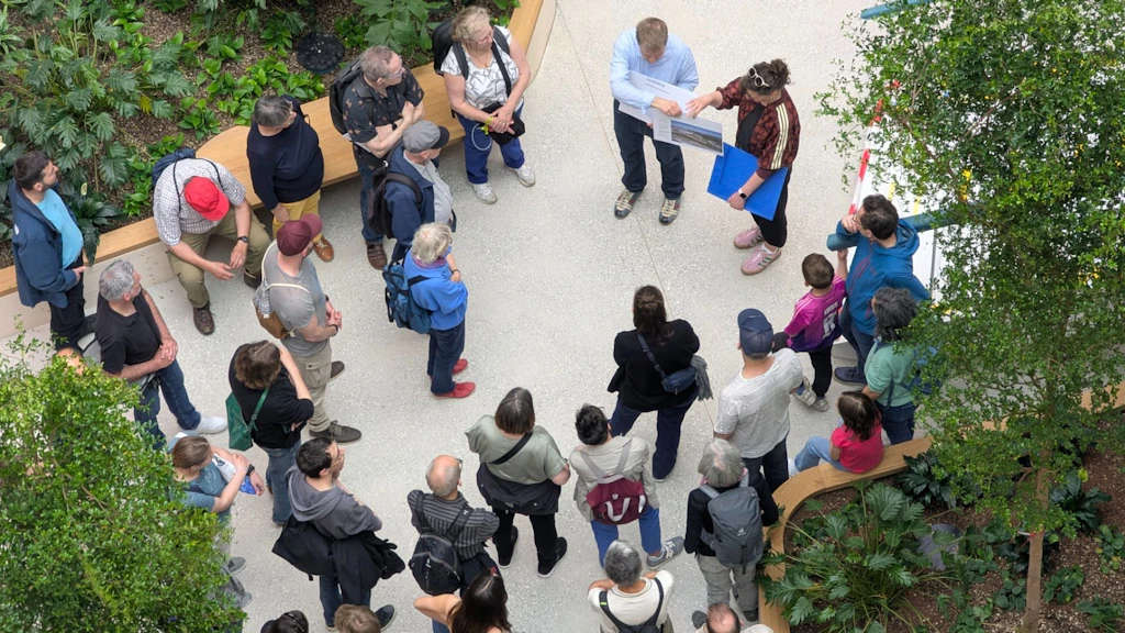 Un groupe de personnes se trouve dans une cour intérieure lumineuse, agrémentée de nombreuses plantes. Elles écoutent deux personnes qui donnent des explications. Cette photo a été prise lors de l'événement Open House Bern.
