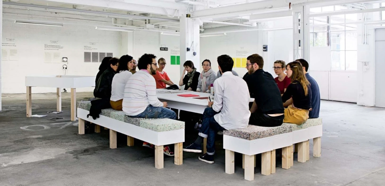 Plusieurs personnes sont assises autour d’une grande table dans un espace d’exposition lumineux et discutent. L’environnement ouvert, avec des panneaux de texte et des installations, montre un échange commun.