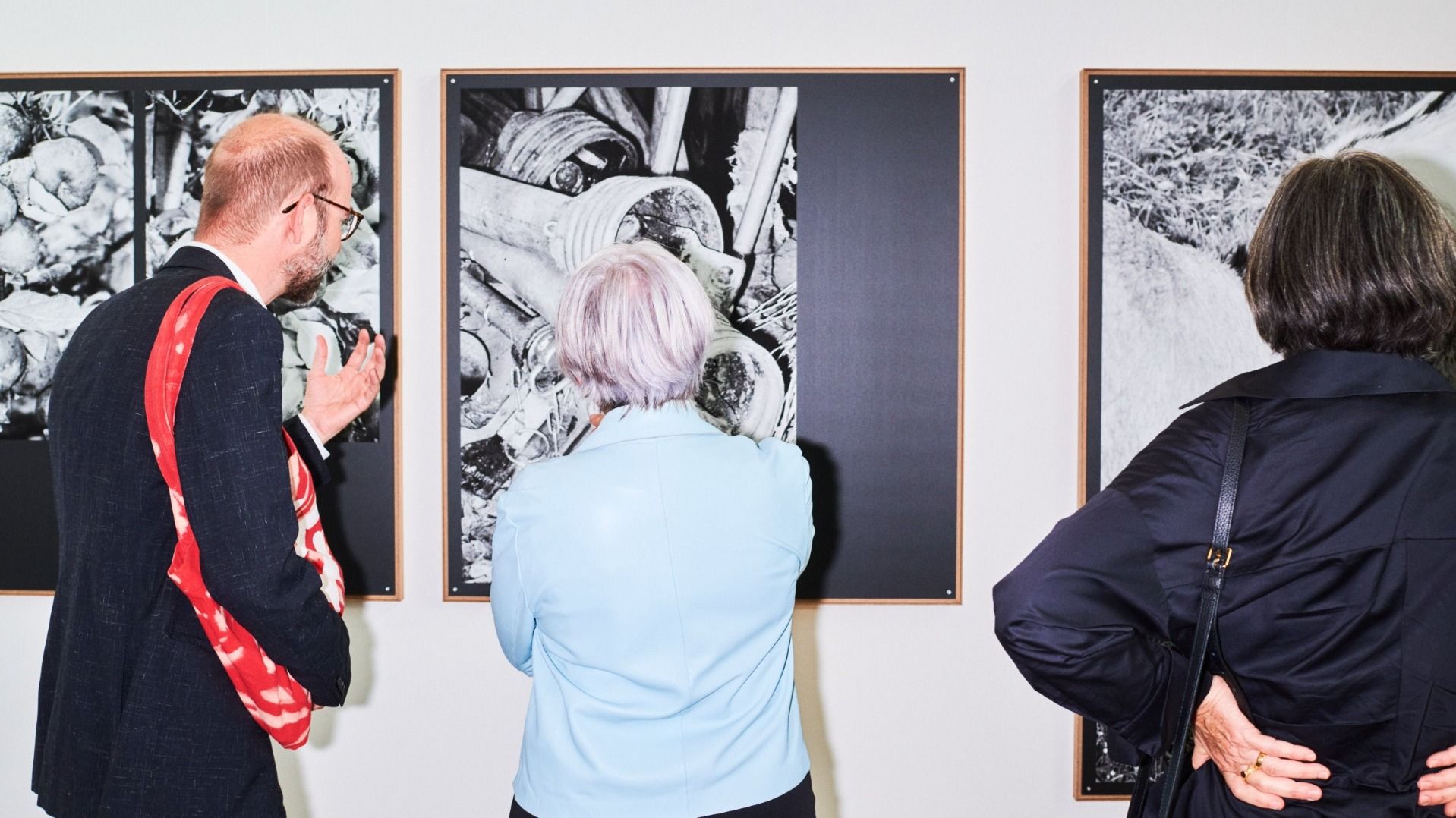 Tre persone osservano opere in bianco e nero in una mostra. Le immagini raffigurano strutture e oggetti dettagliati, incorniciati su una parete bianca.